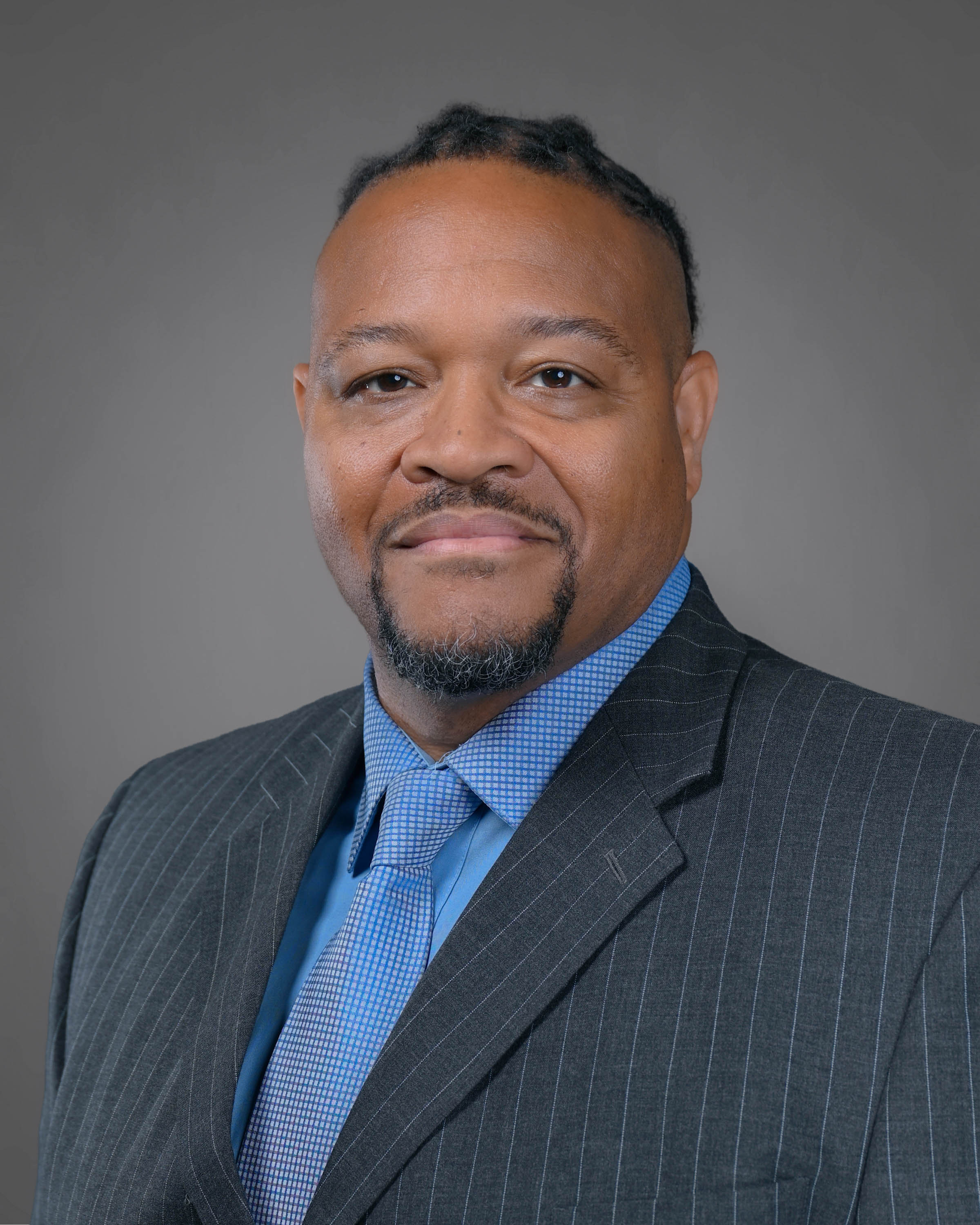 Smiling man with dark curly hair and a goatee in a grey pin striped suit with a blue shirt